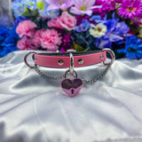 Pink collar with a heart-shaped lock charm on a white surface with flowers in the background