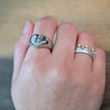 A close-up of a hand wearing two silver rings, with a larger signet-style ring on the middle finger and a smaller crown-like ring on the ring finger, set against a blurred wooden background.